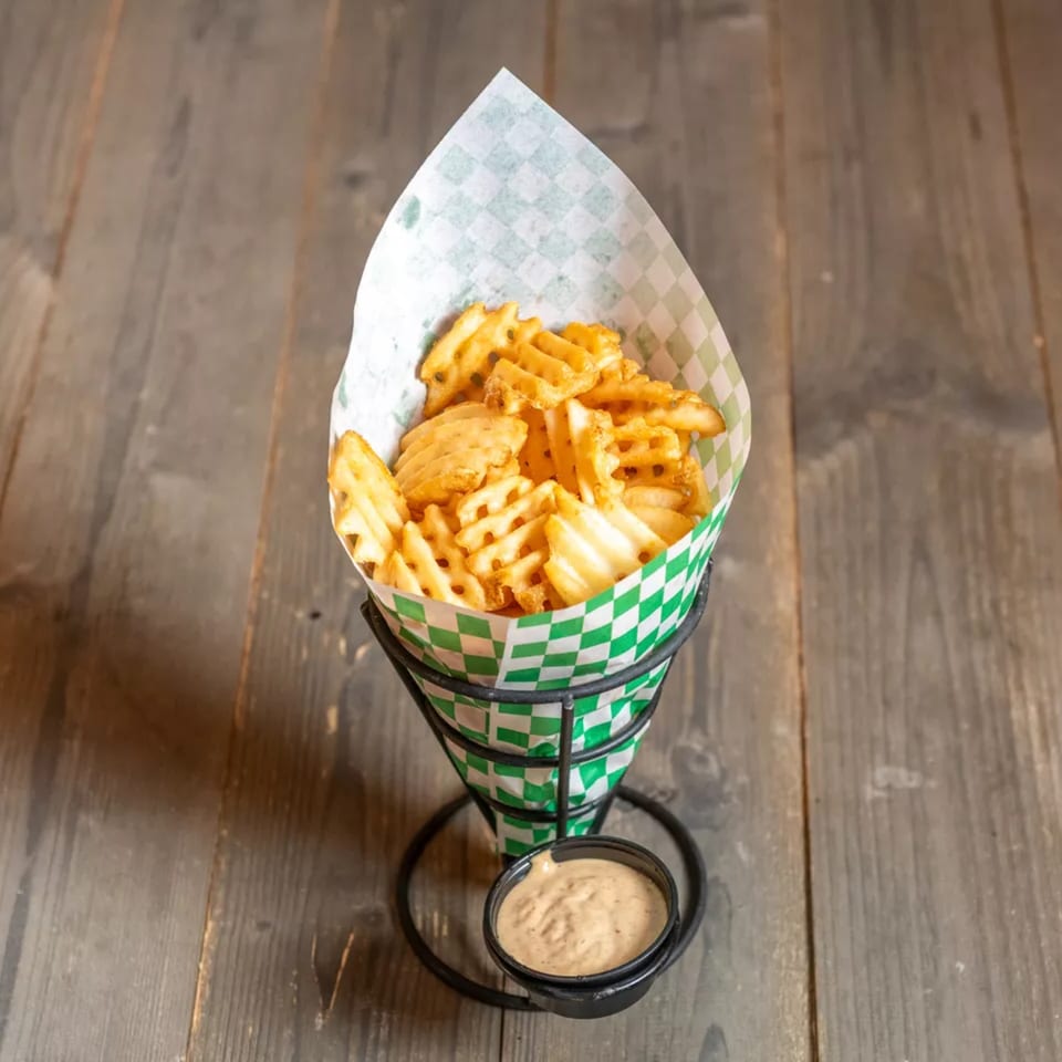 Crispy waffle fries in a green and white checkered paper cone, with a side of dipping sauce.