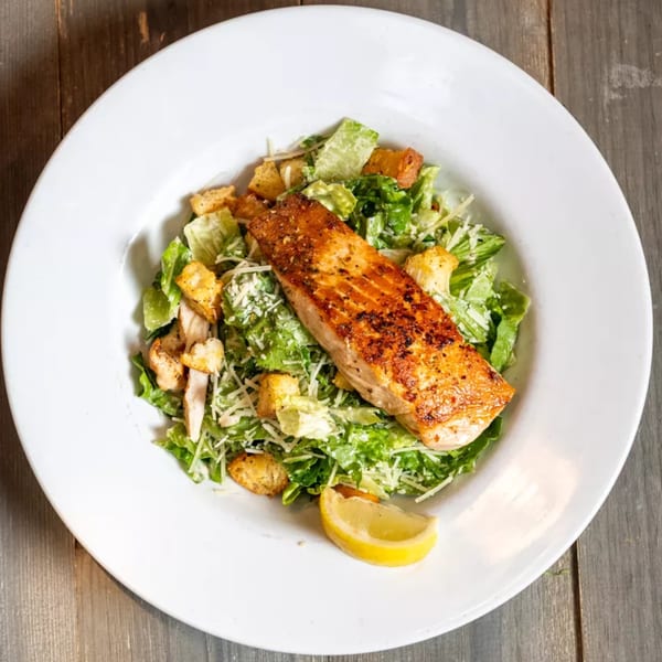 Grilled salmon Caesar salad with croutons, shredded cheese, and a lemon wedge on a white plate.