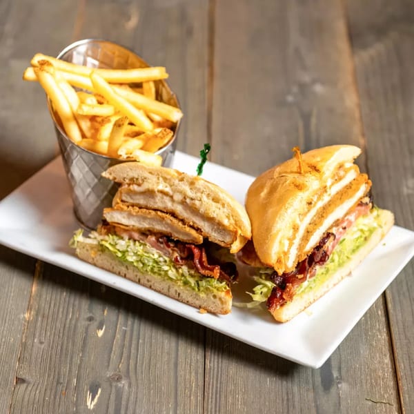 A chicken sandwich with bacon, lettuce, and tomato, served with a metal cup of french fries.