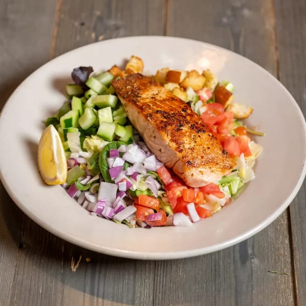 Grilled salmon fillet on a fresh salad with cucumbers, tomatoes, red onions, and a lemon wedge.